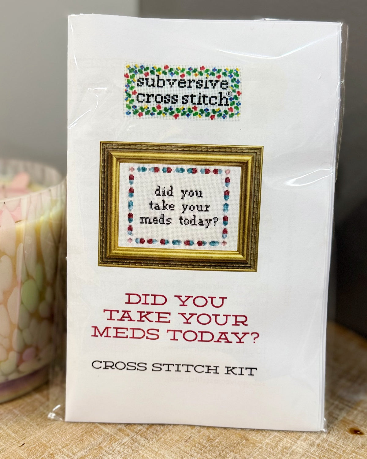 A packaged cross stitch kit labeled 'subversive cross stitch' with the phrase 'did you take your meds today?' shown on the front, photographed on a tabletop next to a candle.