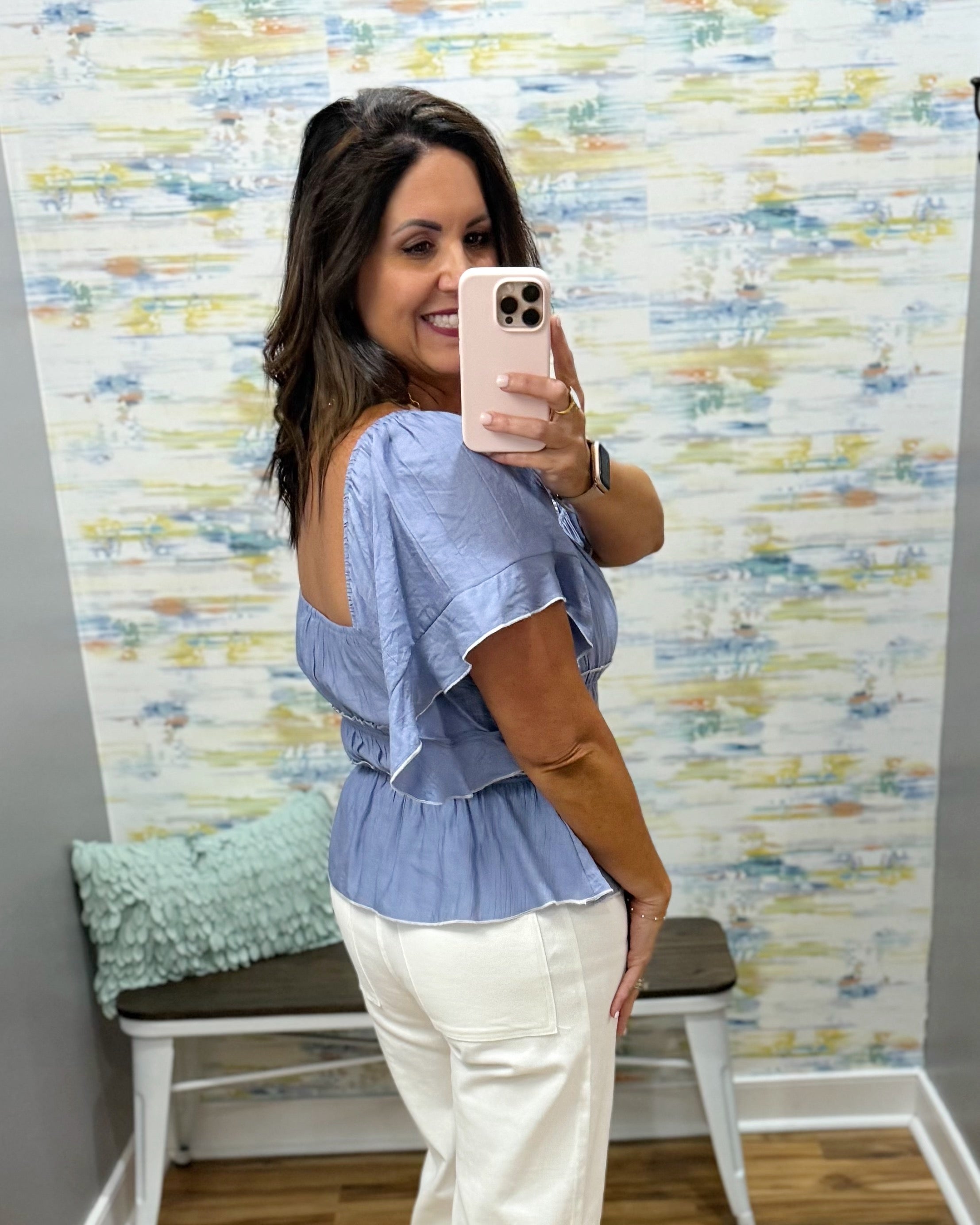 Woman taking a mirror selfie wearing a blue top and white pants in front of a colorful wall.
