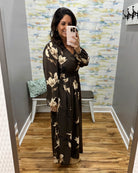 A woman takes a mirror selfie in a fitting room wearing a long-sleeve, V-neck maxi dress with a dark brown base and a beige floral print. The background features a gray wall, a bench, and a patterned wallpaper.