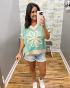Woman taking a mirror selfie wearing a green and beige floral-patterned top and denim shorts.