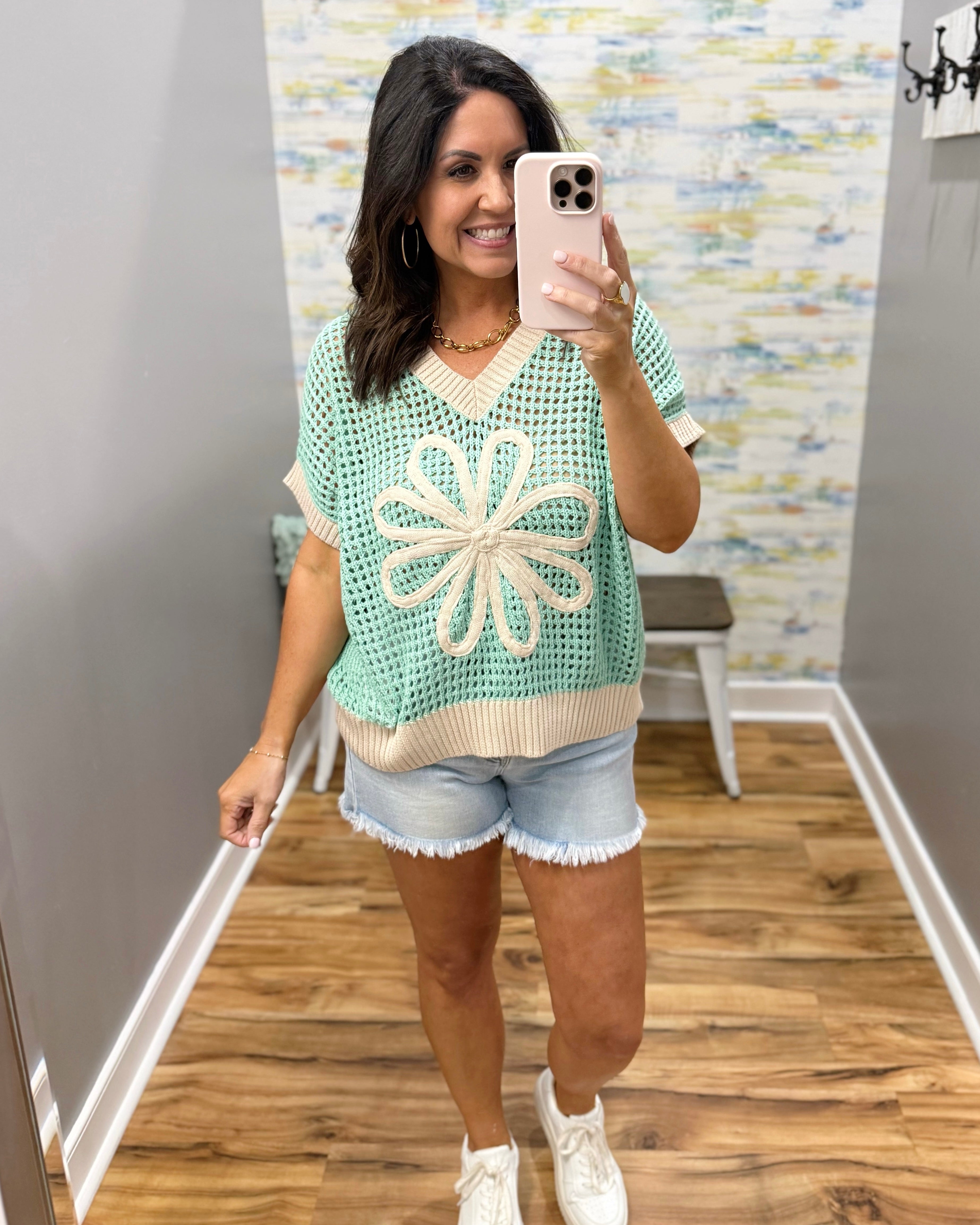 Woman taking a mirror selfie wearing a green and beige floral-patterned top and denim shorts.