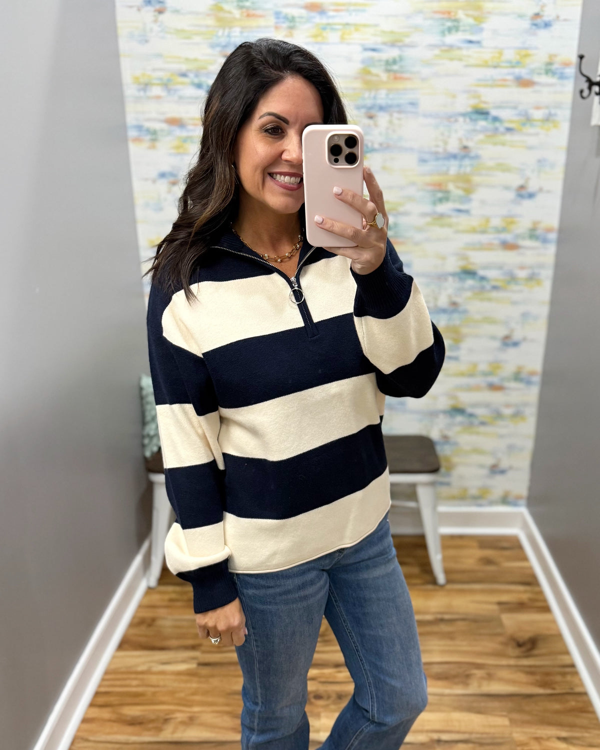 A woman takes a mirror selfie wearing a navy blue and cream striped quarter-zip pullover sweater with blue jeans, standing in a fitting room with a patterned wall and wood floor.