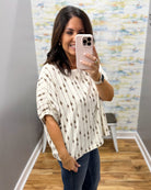 A woman taking a mirror selfie in a boutique, wearing a cream-colored short-sleeve knit top with vertical brown dotted lines and small diamond motifs, paired with dark blue jeans.