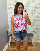 Woman taking a mirror selfie wearing a red floral top and denim shorts in a room with a textured wall.