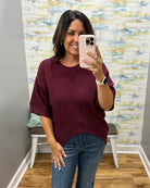 A woman takes a mirror selfie wearing a short-sleeve crewneck knit sweater in burgundy and blue jeans, standing in front of a patterned wall.
