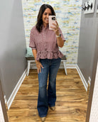 A woman takes a mirror selfie wearing a red and white gingham short-sleeve blouse with a ruffled hem and dark blue flare jeans in a home setting with wood floors and a patterned accent wall.