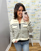 A woman takes a mirror selfie wearing a cream zip-up jacket with black horizontal stripes, paired with a black top and blue jeans, in front of a patterned wallpaper background.