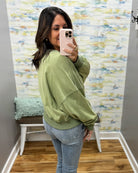 A woman takes a mirror selfie in a fitting room wearing an oversized sage green sweatshirt and light blue jeans.
