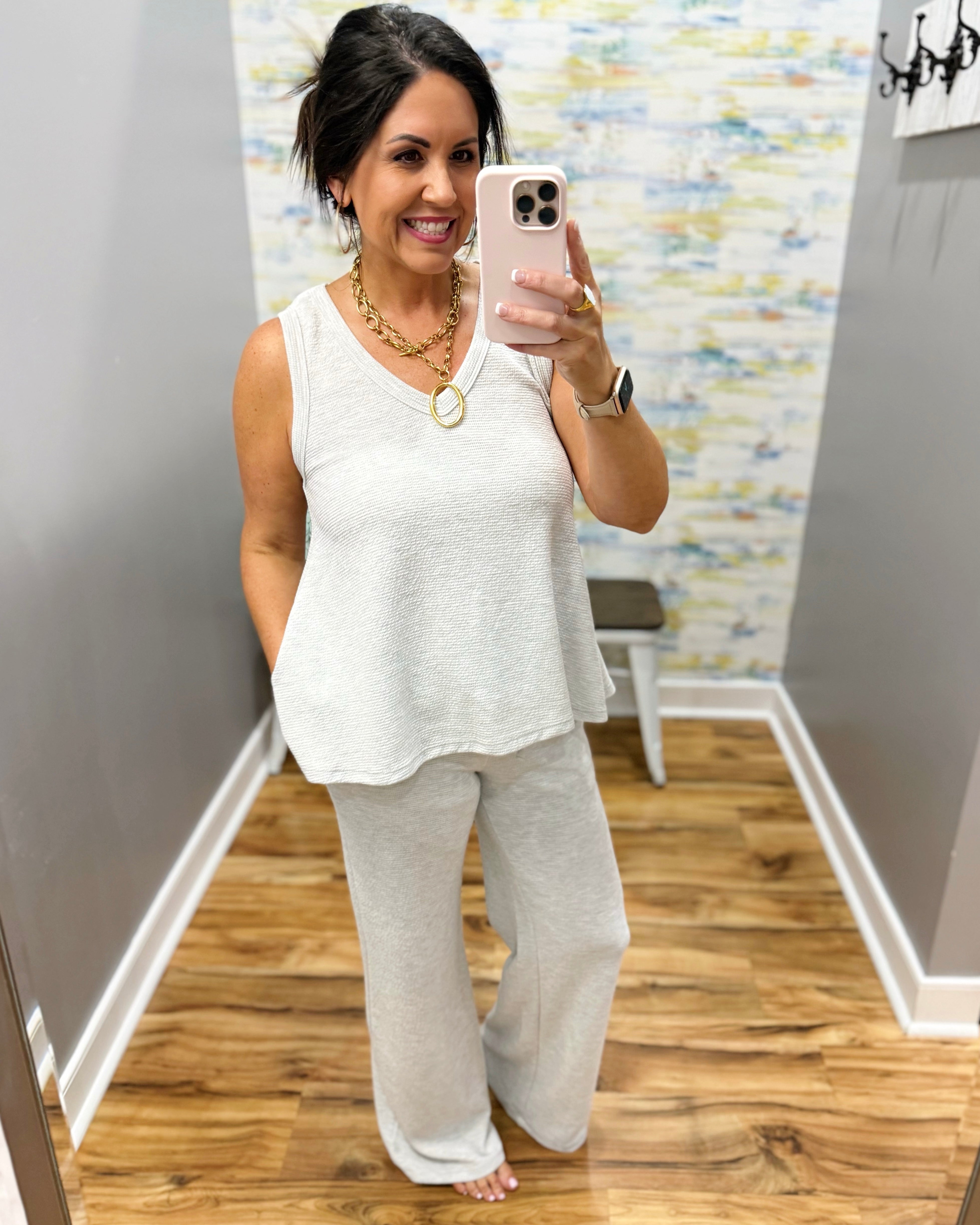 Woman taking a mirror selfie wearing a white sleeveless top and light gray pants in a room with a colorful wall.