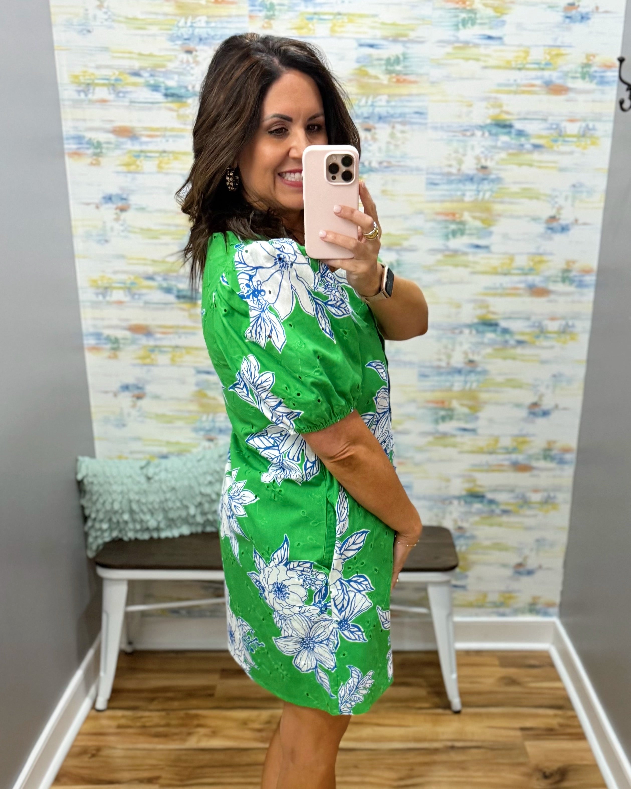 Woman taking a mirror selfie wearing a green floral dress in a room with a textured wall.