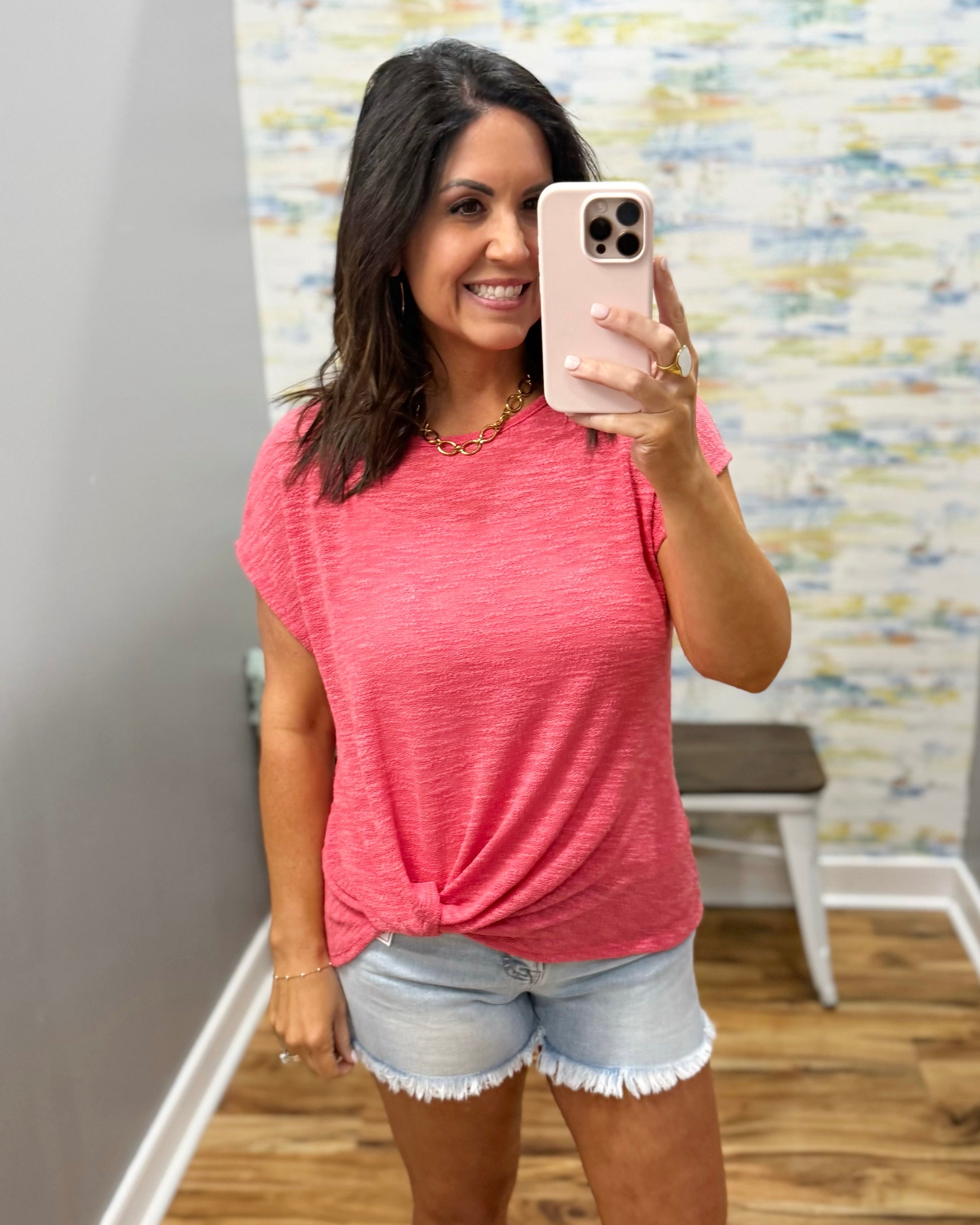 Woman taking a mirror selfie wearing a pink t-shirt and denim shorts in a room with a colorful wall.