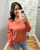 A woman takes a mirror selfie wearing a long-sleeve V-neck knit sweater in a muted terracotta or burnt orange color, paired with blue jeans.