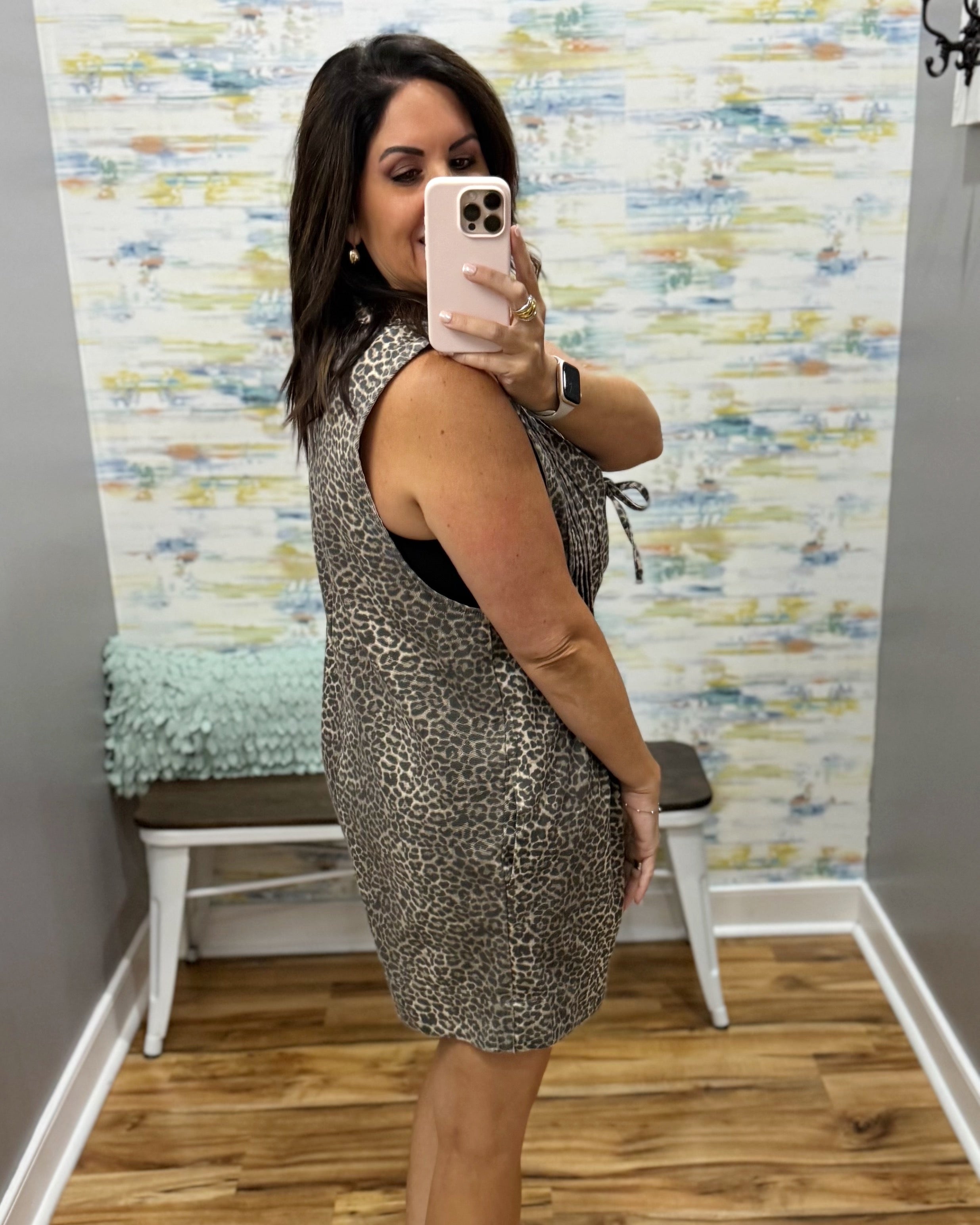 A woman takes a mirror selfie in a boutique showing the side profile of a sleeveless leopard-print mini dress. The dress has a brown and beige animal pattern over a black underlayer.