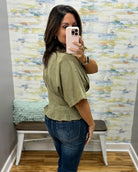 A woman takes a mirror selfie showing the back of an olive green short-sleeve peplum top paired with blue jeans, set against a patterned wallpaper background.