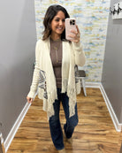 A woman takes a mirror selfie in a fitting room wearing a cream open-front knit cardigan with fringe and geometric patterned details on the sleeves, layered over a brown top and dark blue jeans.