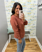 A woman takes a mirror selfie wearing a textured, long-sleeve crewneck sweater in a dusty terracotta color, paired with light blue jeans. The setting is a home fitting room with gray walls, a wooden floor, and a colorful abstract-patterned wallpaper.