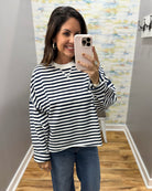 A woman takes a mirror selfie wearing a navy and white horizontally striped long-sleeve top with blue jeans, holding a light pink smartphone.