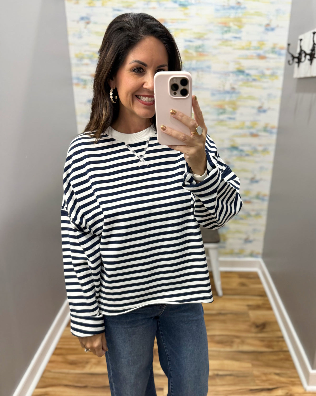 A woman takes a mirror selfie wearing a navy and white horizontally striped long-sleeve top with blue jeans, holding a light pink smartphone.
