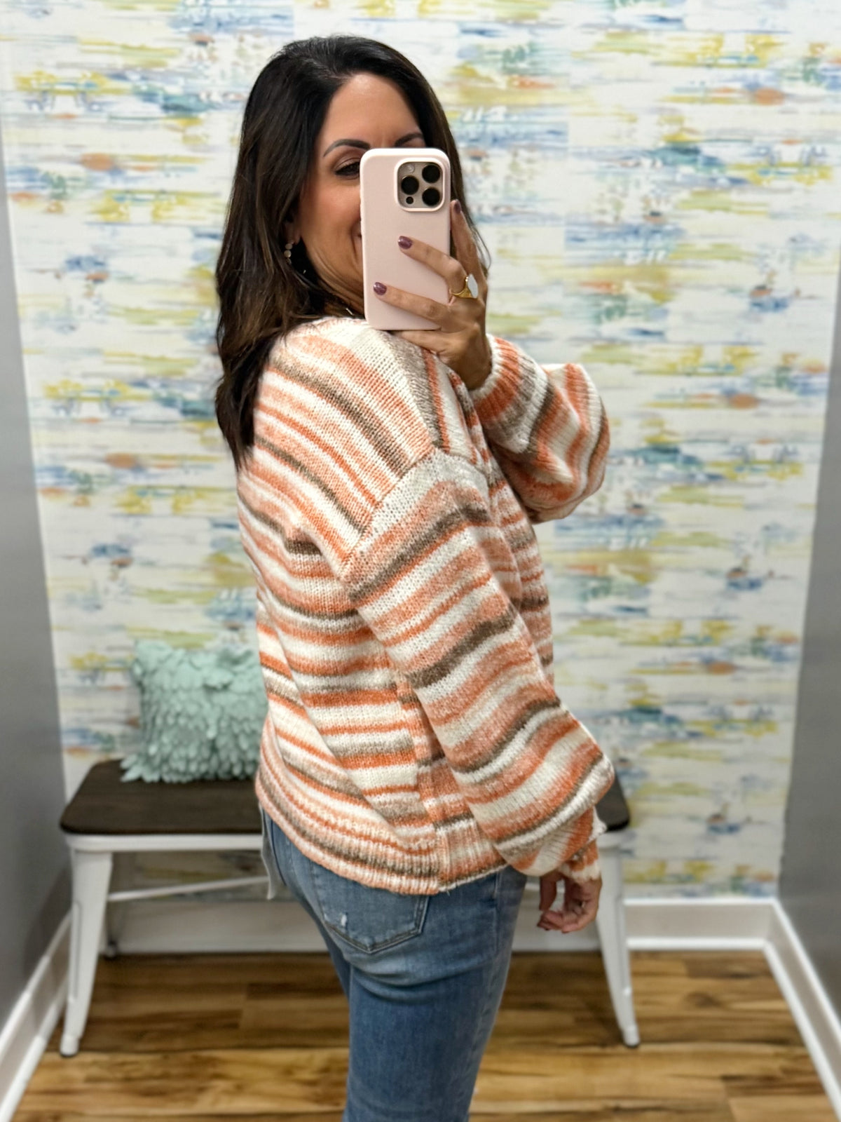 A woman takes a mirror selfie wearing a fuzzy striped knit sweater in peach, cream, and taupe, paired with blue jeans, in front of a patterned wallpaper wall.