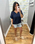Woman taking a mirror selfie wearing a navy blue top and denim shorts in a room with a textured wall.