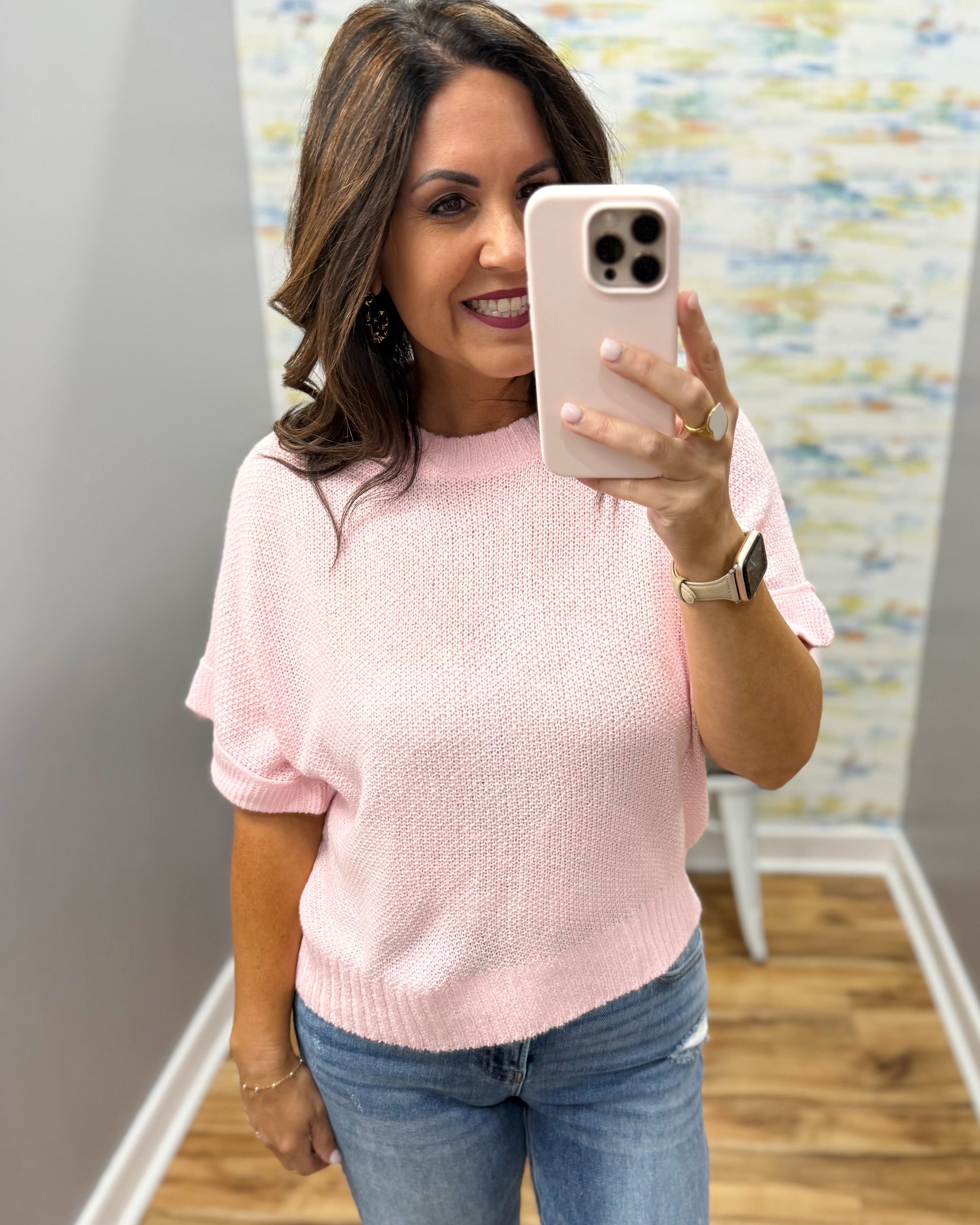 Woman taking a mirror selfie wearing a pink sweater and blue jeans in a room with a colorful wall.