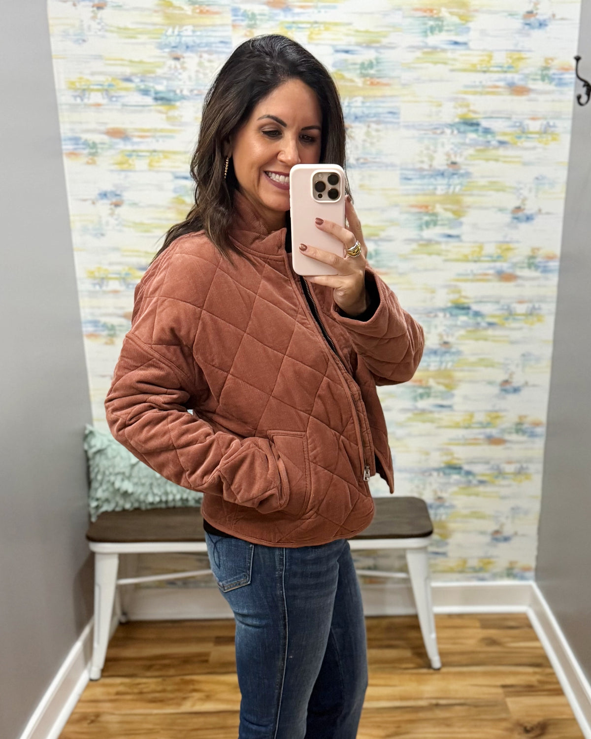 A woman takes a mirror selfie wearing a rust-brown diamond-quilted jacket and blue jeans in a fitting room with a patterned wallpaper background.