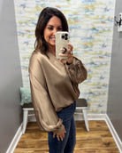 A woman takes a mirror selfie wearing a shiny taupe satin long-sleeve blouse and blue jeans in a home setting with a patterned wallpaper background.