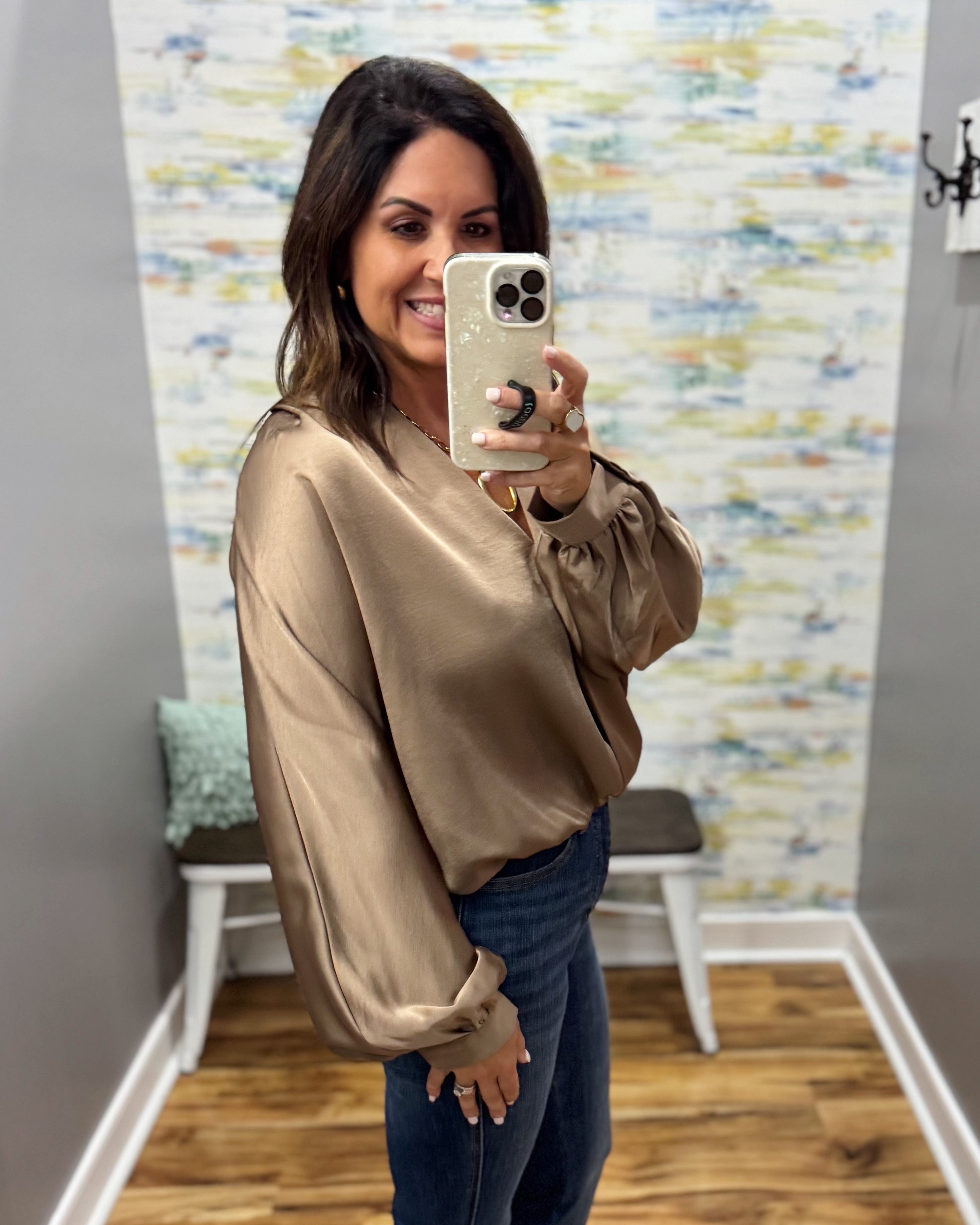A woman takes a mirror selfie wearing a shiny taupe satin long-sleeve blouse and blue jeans in a home setting with a patterned wallpaper background.