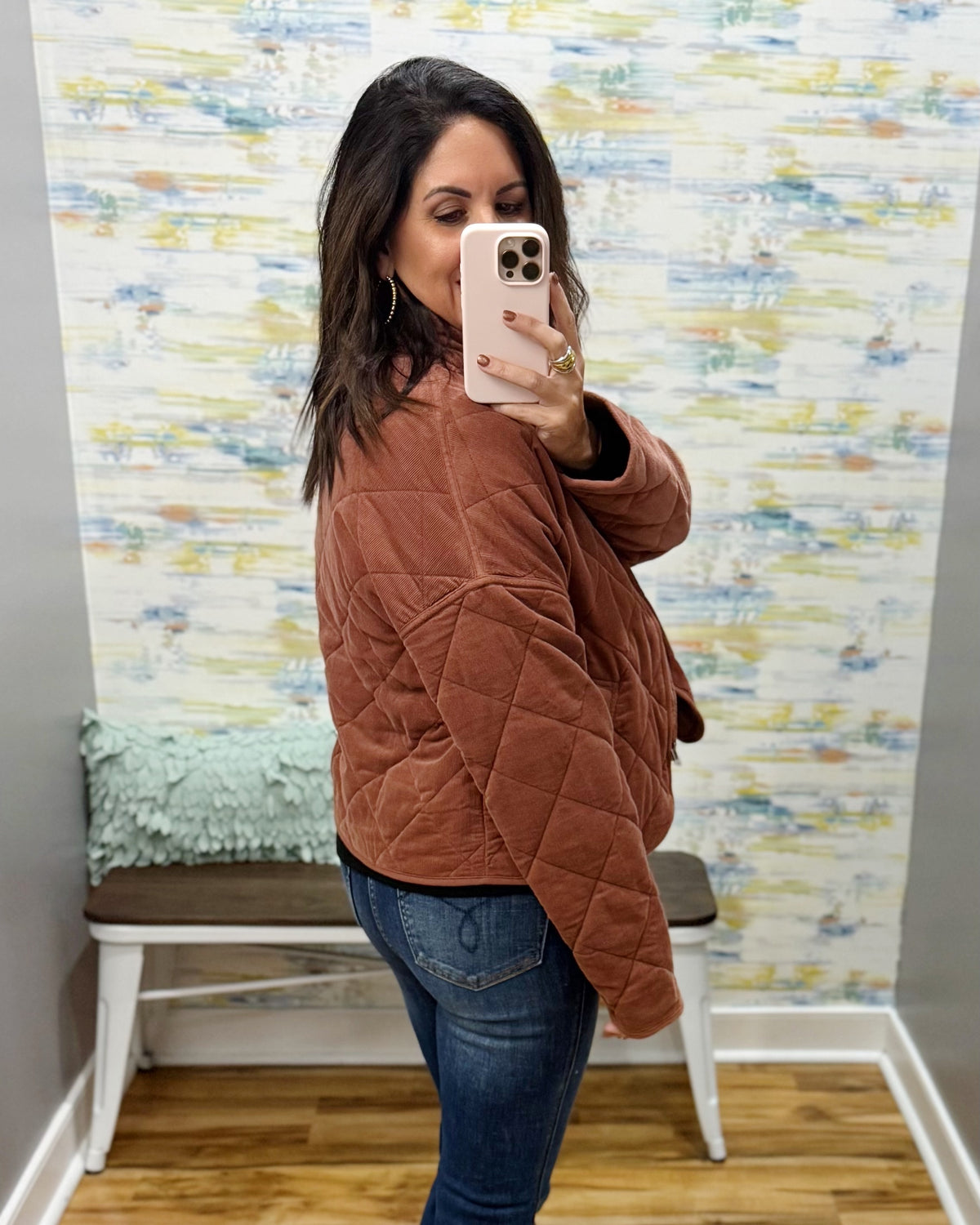 A woman takes a mirror selfie wearing a rust-brown diamond-quilted jacket and blue jeans in front of a patterned wallpaper background.