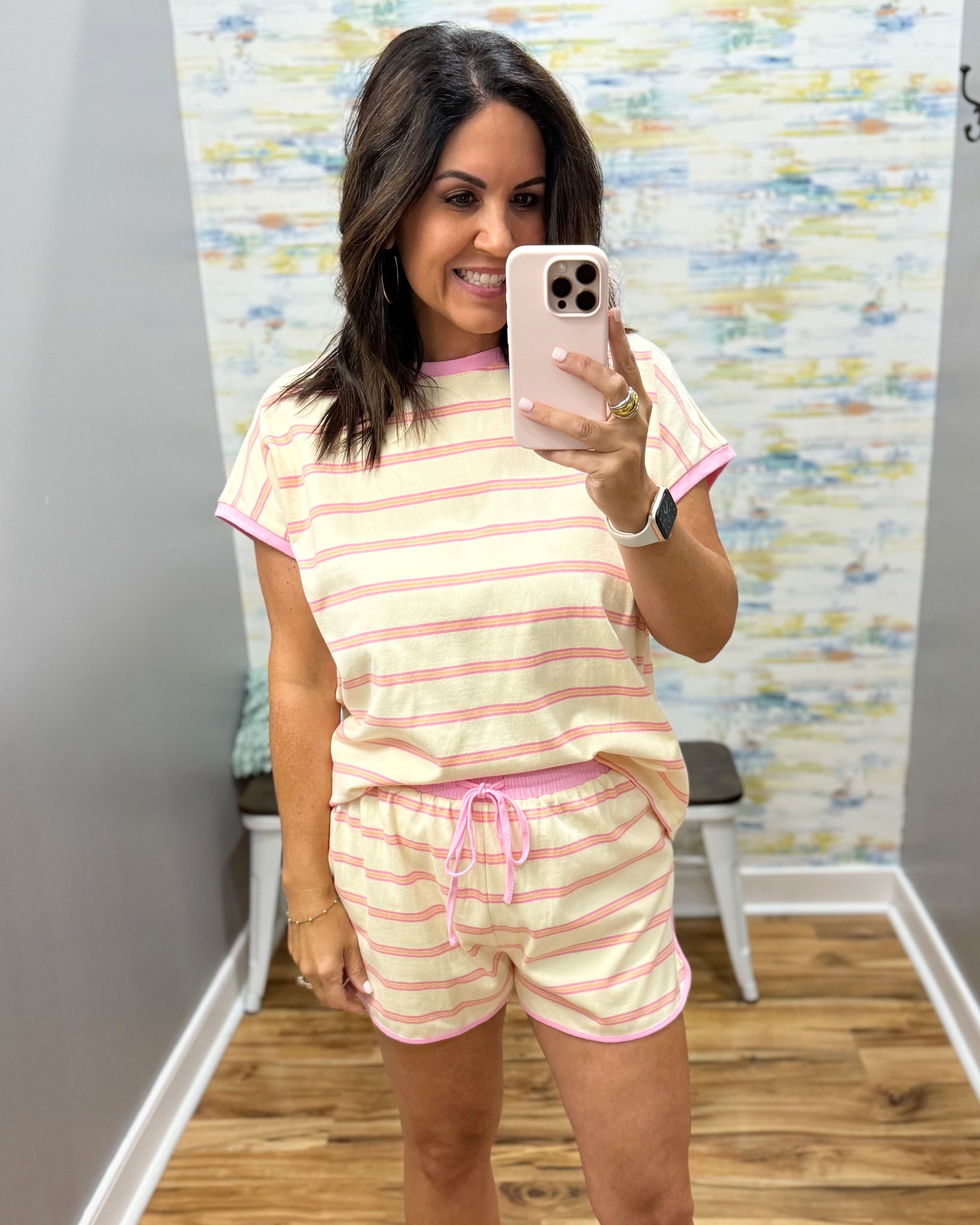 A woman takes a mirror selfie wearing a matching two-piece loungewear set with pink and cream horizontal stripes, featuring a short-sleeve t-shirt and shorts with a pink drawstring.