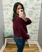 A woman taking a mirror selfie in a fitting room, wearing a burgundy long-sleeve bubble-sleeve top and blue jeans.