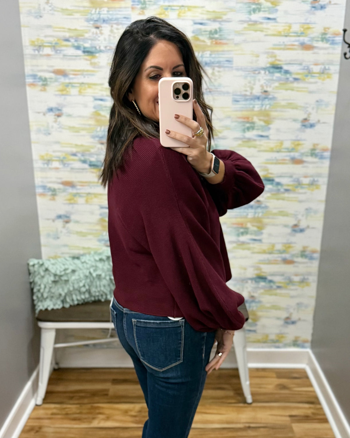 A woman taking a mirror selfie in a fitting room, wearing a burgundy long-sleeve bubble-sleeve top and blue jeans.
