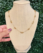 A gold chain necklace with small decorative bead-like links, displayed on a beige fabric jewelry bust. A hand with purple nail polish is holding the left side of the bust, and a green leafy background is visible behind it.