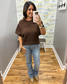 A woman takes a mirror selfie wearing a brown short-sleeve crewneck knit top, light-wash blue jeans, and beige ankle boots in a home fitting room.