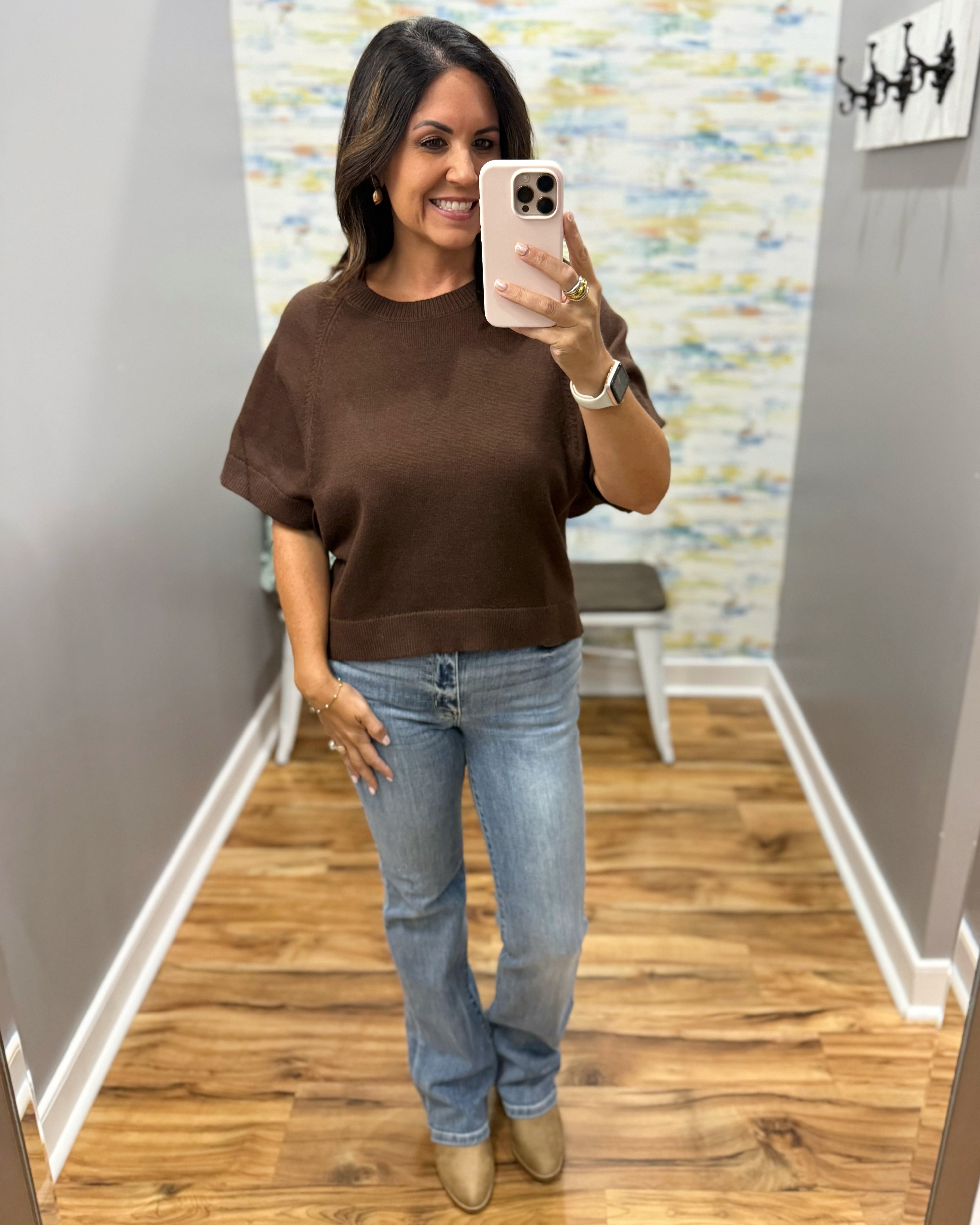 A woman takes a mirror selfie wearing a brown short-sleeve crewneck knit top, light-wash blue jeans, and beige ankle boots in a home fitting room.