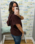 A woman takes a mirror selfie wearing a short-sleeve, loose-fitting knit top in a deep burgundy or maroon color, paired with blue jeans. She is standing in front of a patterned wallpaper background with a white bench and a mint green pillow visible.