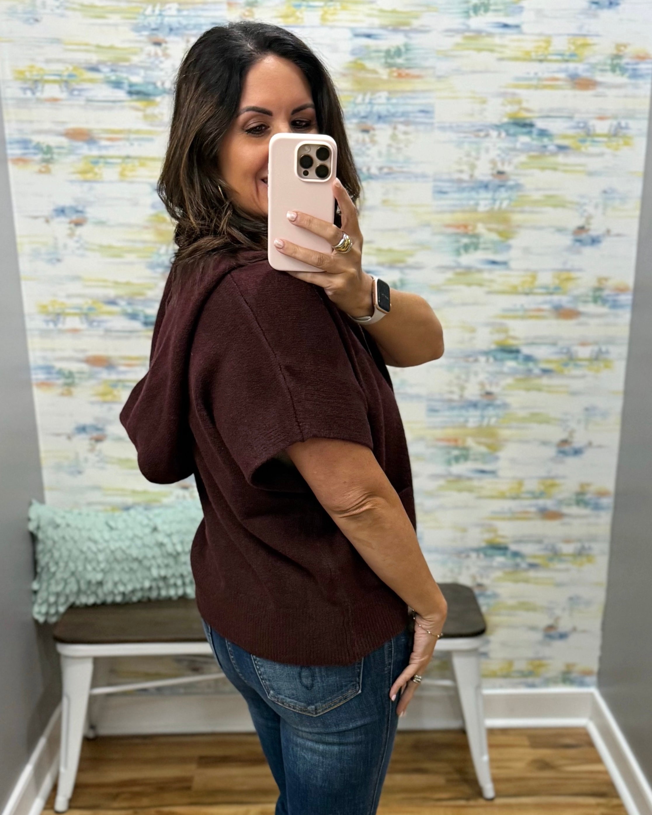 A woman takes a mirror selfie wearing a short-sleeve, loose-fitting knit top in a deep burgundy or maroon color, paired with blue jeans. She is standing in front of a patterned wallpaper background with a white bench and a mint green pillow visible.