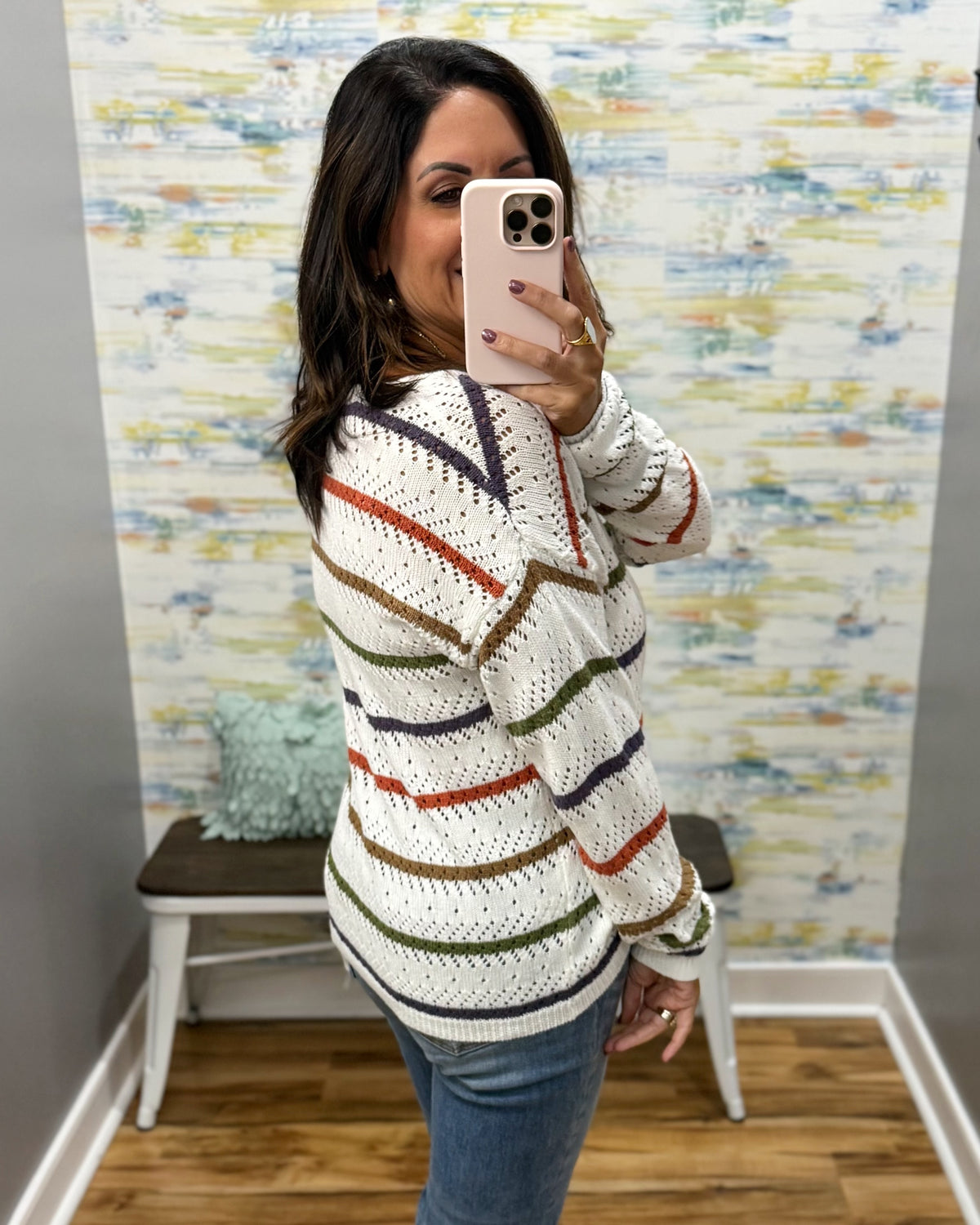 A woman takes a mirror selfie wearing a white knit sweater with horizontal stripes in orange, green, brown, and navy, paired with blue jeans.