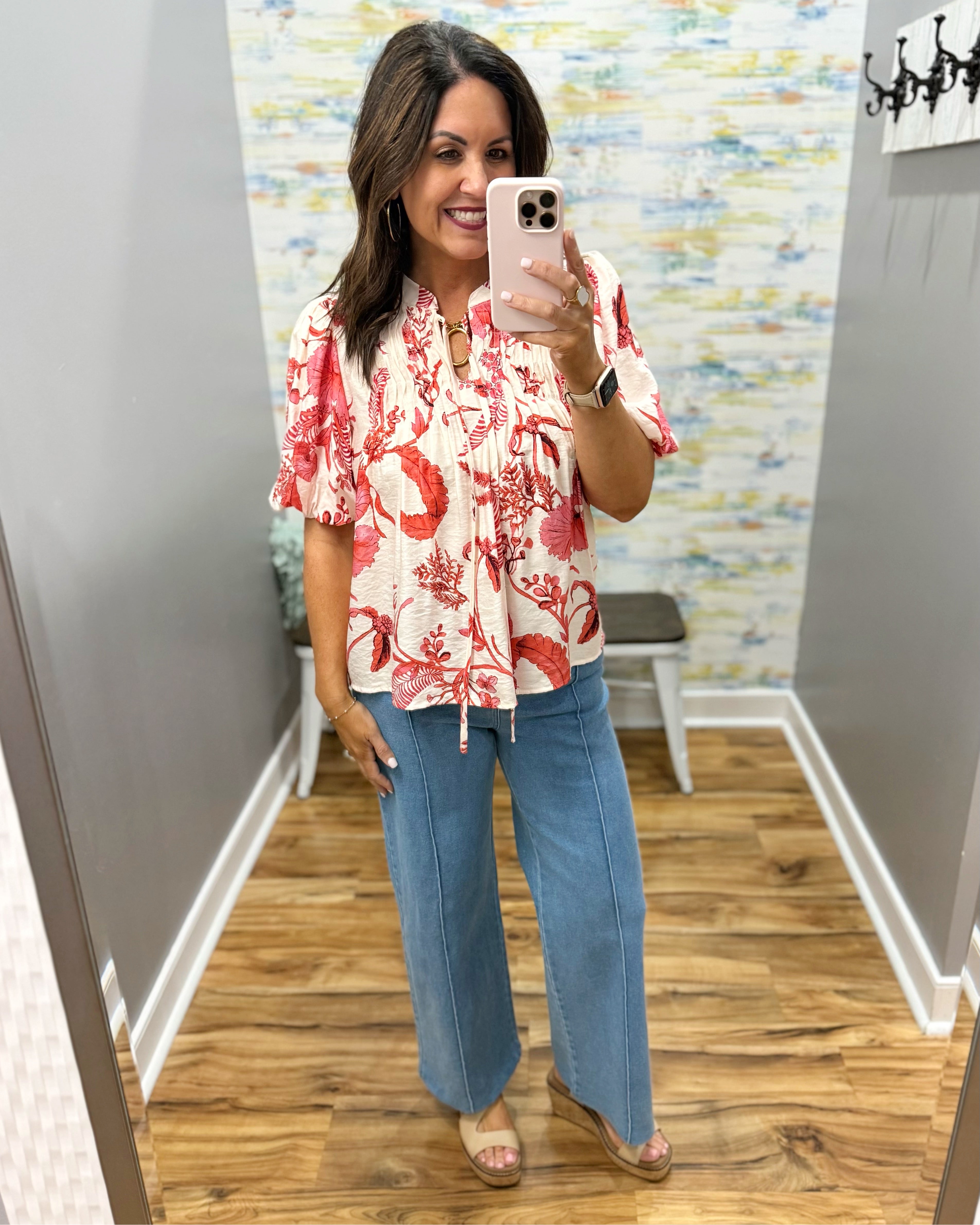 Woman taking a mirror selfie wearing a floral top and blue jeans in a room with patterned wallpaper.