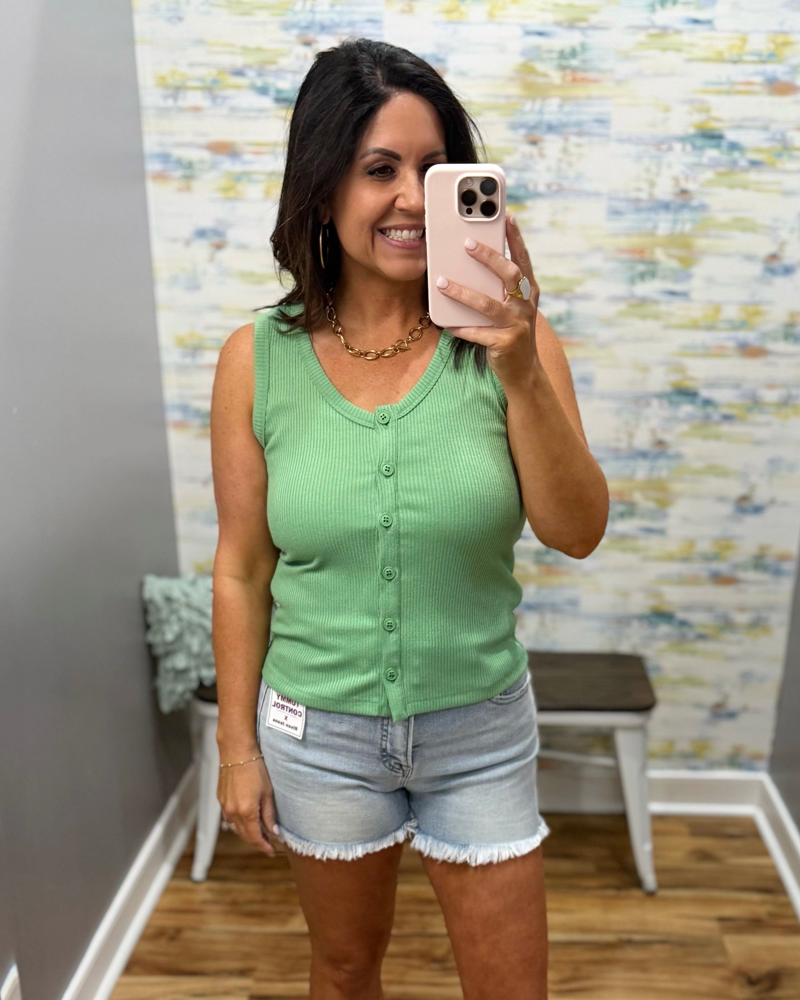 Woman taking a mirror selfie wearing a green sleeveless top and denim shorts in a room with a textured wall.