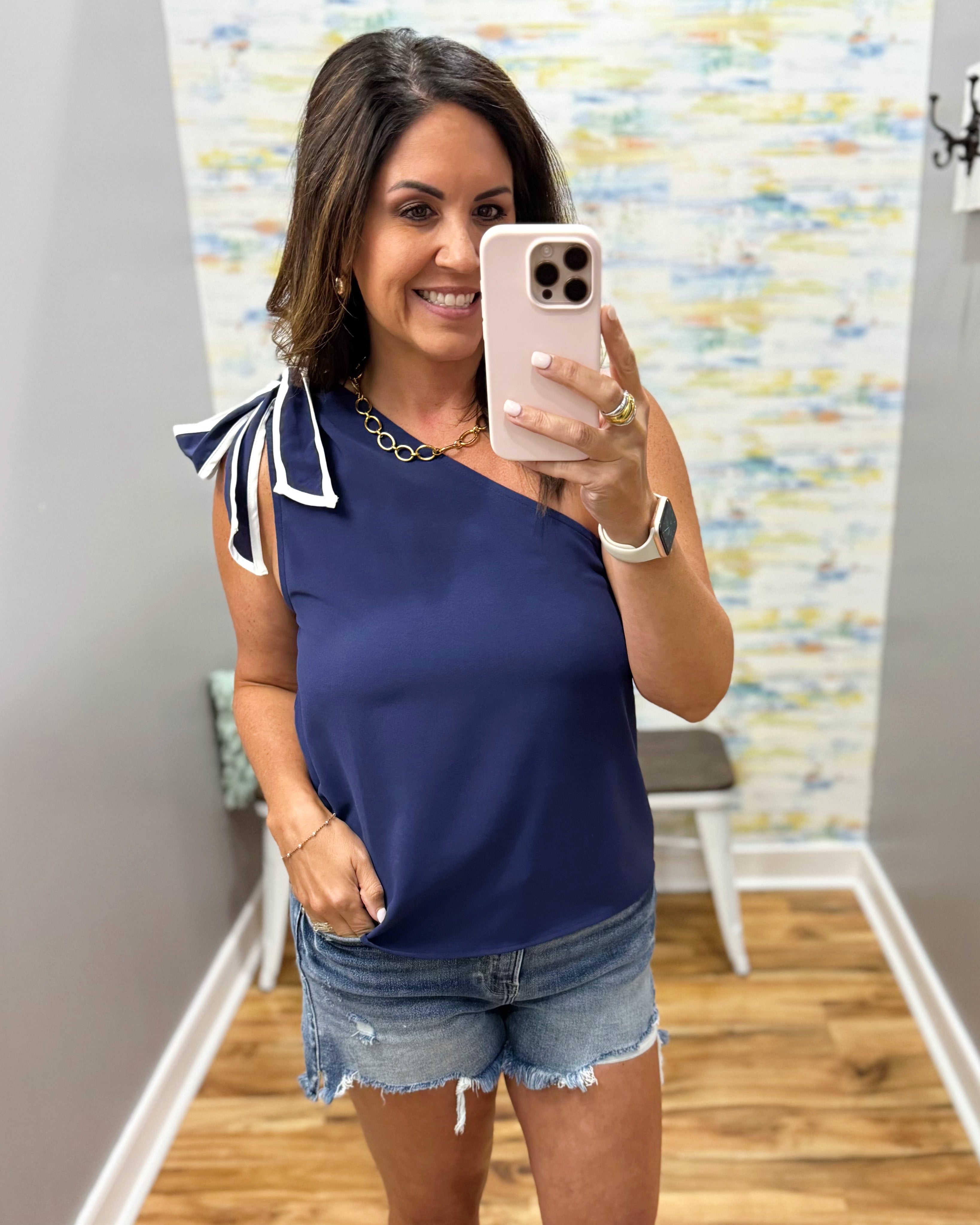 Woman taking a mirror selfie wearing a navy blue top with white trim and denim shorts.