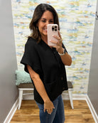 A woman taking a mirror selfie in a fitting room wearing a black short-sleeve button-front top and blue jeans.