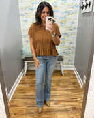A woman takes a mirror selfie in a fitting room wearing a rust-brown short-sleeve ruffled top, light-wash blue jeans, and tan boots.