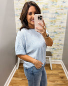 Woman taking a mirror selfie wearing a light blue sweater and jeans in a room with patterned wallpaper.