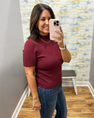 A woman taking a mirror selfie in a fitting room wearing a short-sleeve ribbed knit top in burgundy and blue jeans.