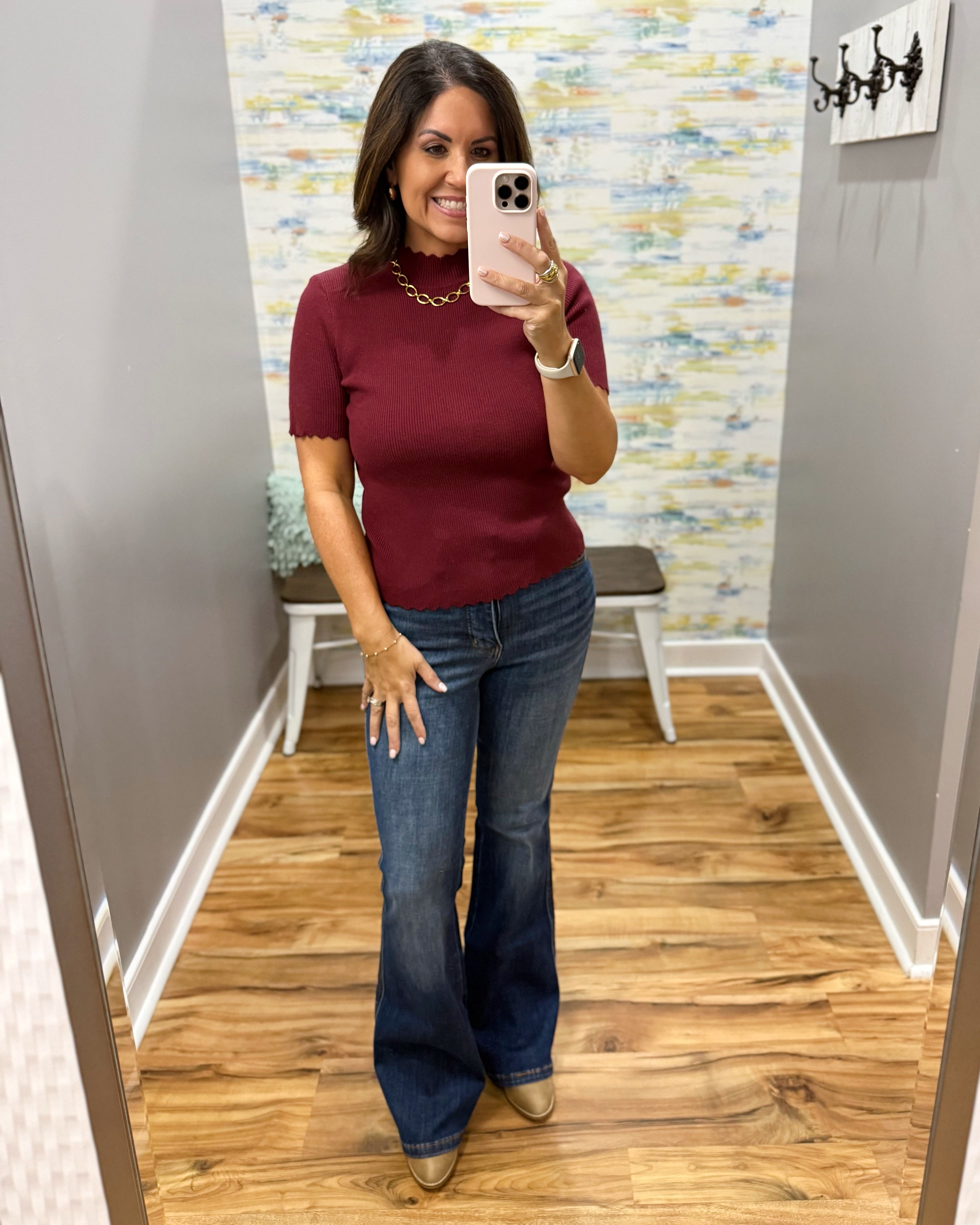 A woman takes a mirror selfie in a fitting room wearing a short-sleeve burgundy knit top and blue flare jeans.