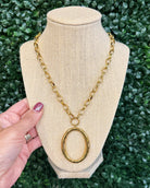 Chansuttpearls Alice gold chain necklace with a large oval pendant displayed on a beige fabric jewelry bust, with a hand holding the left side against a green leaf background.