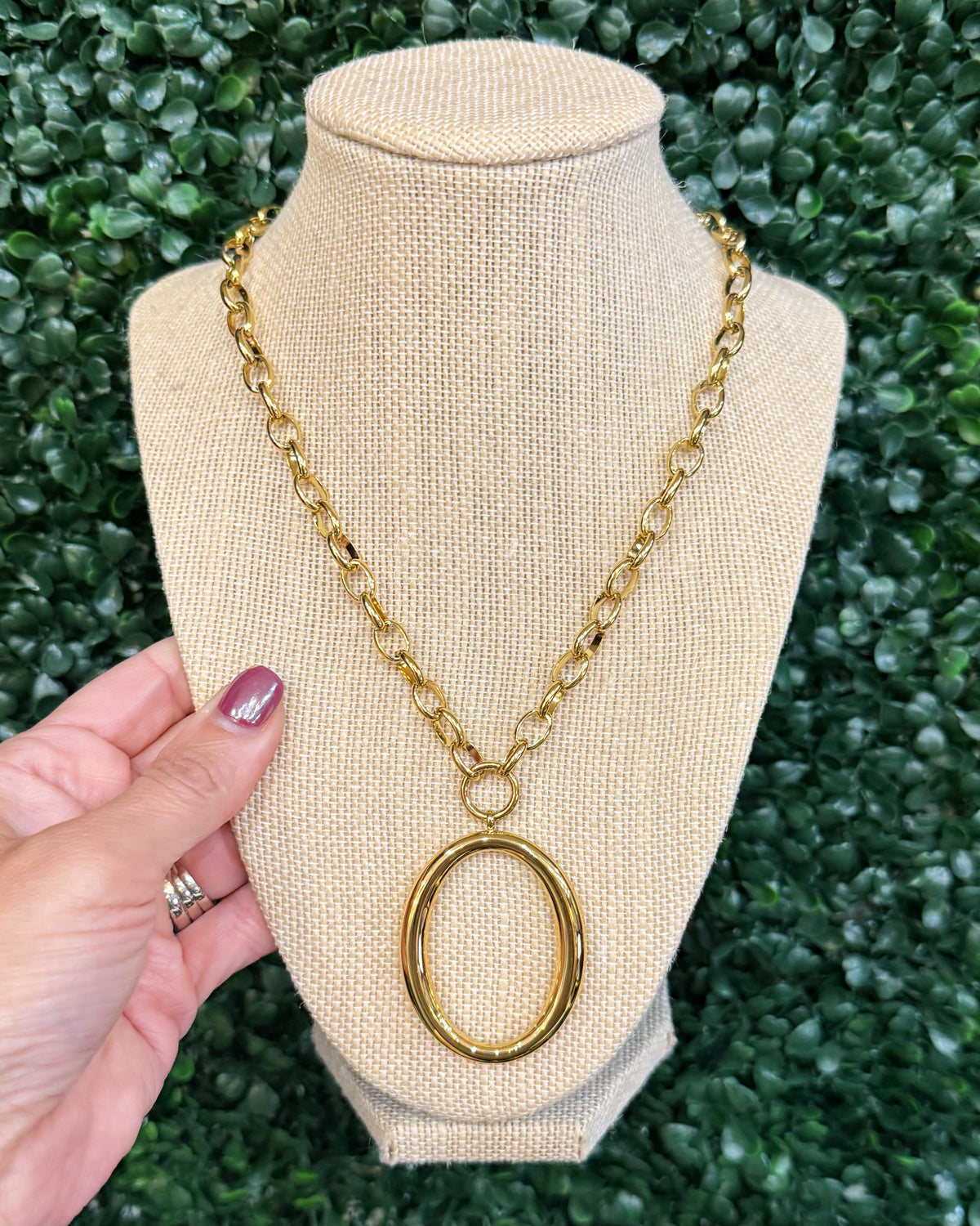 Chansuttpearls Alice gold chain necklace with a large oval pendant displayed on a beige fabric jewelry bust, with a hand holding the left side against a green leaf background.