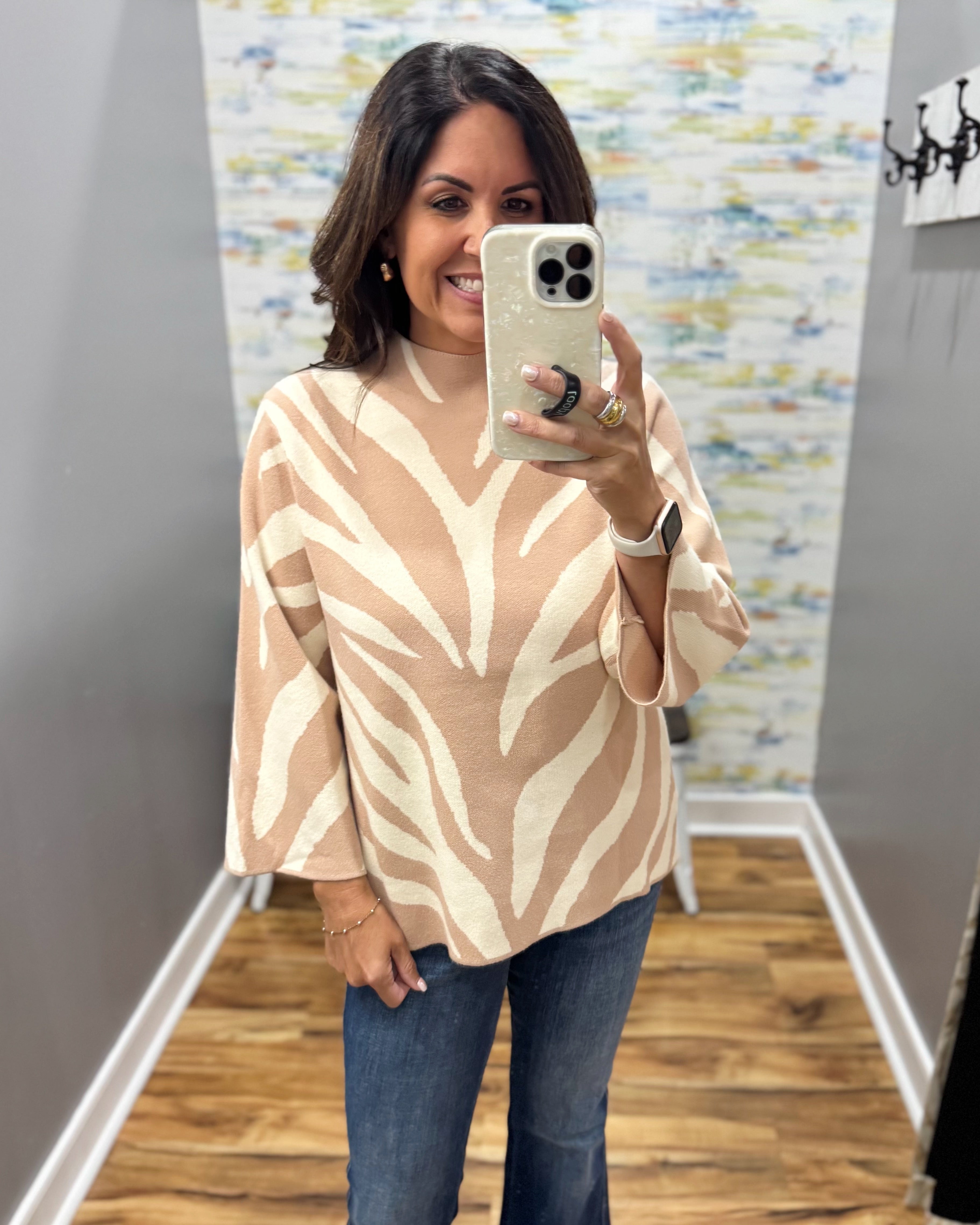 A woman takes a mirror selfie wearing a beige and cream leaf-patterned mock-neck top with wide three-quarter sleeves, paired with blue jeans.