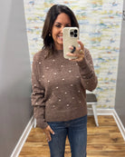 A woman takes a mirror selfie wearing a brown knit sweater with small white polka dots and blue jeans in a fitting room with wood floors and a patterned wall.
